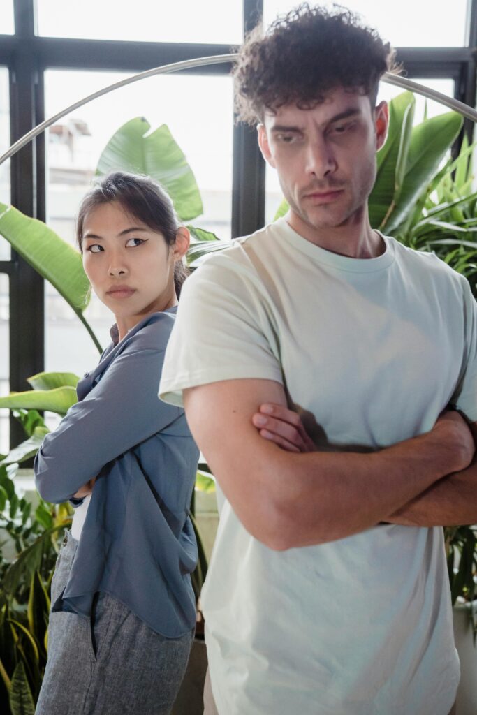 Couple standing back to back with crossed arms and tense expressions, surrounded by indoor plants.